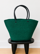 Load image into Gallery viewer, A dark green woven straw tote bag with black rope handles, displayed on a black wooden stool against a white wall.
