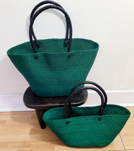 Load image into Gallery viewer, Two woven raffia tote bags in forest green with black handles, displayed on a light wood floor against a white wall. The larger bag stands on a small dark wooden stool, and the smaller bag sits in front of it.
